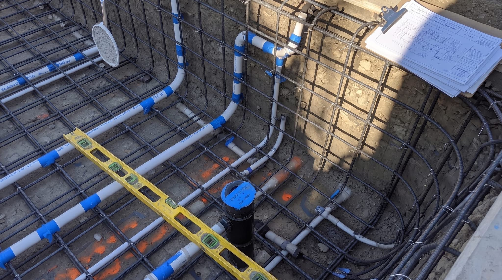 Tight angle close-up inside an excavated pool pit. A grid of freshly tied black rebar fills the frame, forming the structural skeleton of the pool shell. PVC hydraulic plumbing lines rough into the deep end and bond beam. A skimmer body and main-drain fitting are set in their final positions, capped with blue tape. Engineered plans on a clipboard at the deck edge are blurred in the background.