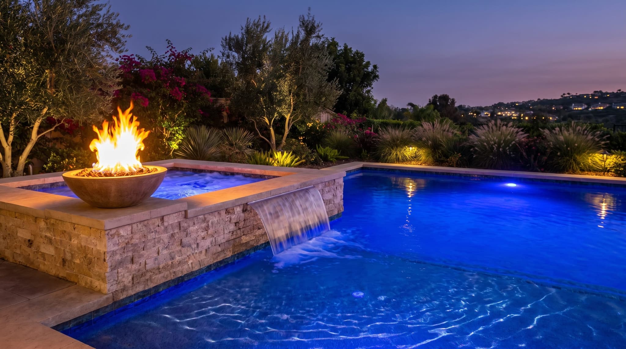 Single warm-stone fire bowl on the corner of a raised spa wall at blue hour with full live flame, sheer-descent waterfall spilling into the main pool below lit deep cobalt from underwater LED, stone wall texture behind