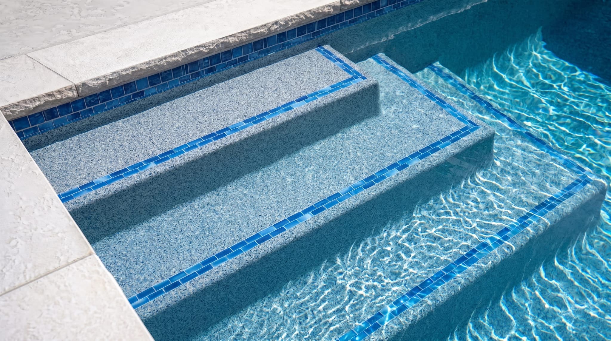 Overhead close-up of a pebble pool interior finish in shallow water with sunlight caustics dancing across the textured surface, varied pebbles in blue-grey, soft brown, and white quartz visible through clear warm-toned water